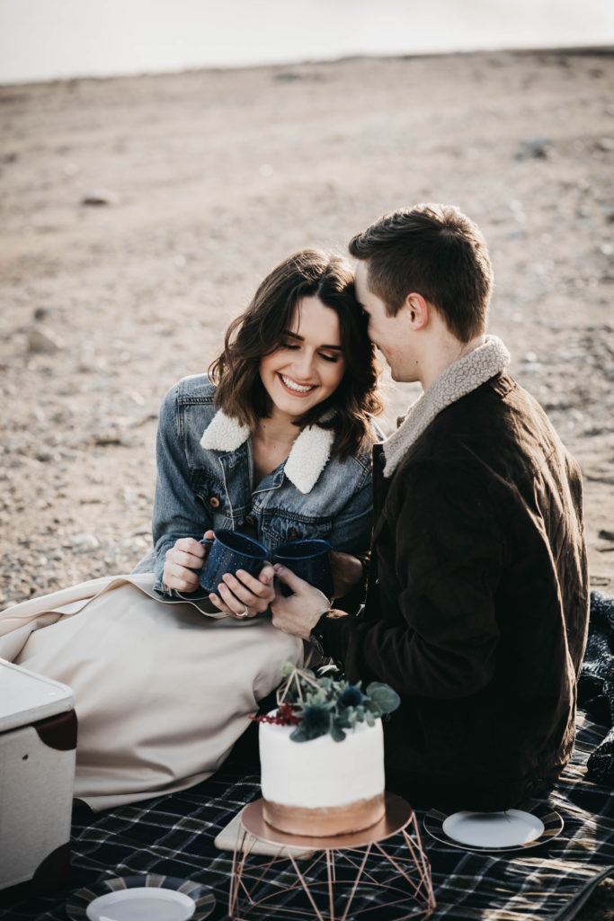 couple tries to get warm together during their lucky peak lake elopement with gimme some sugar elopement cake in front of them