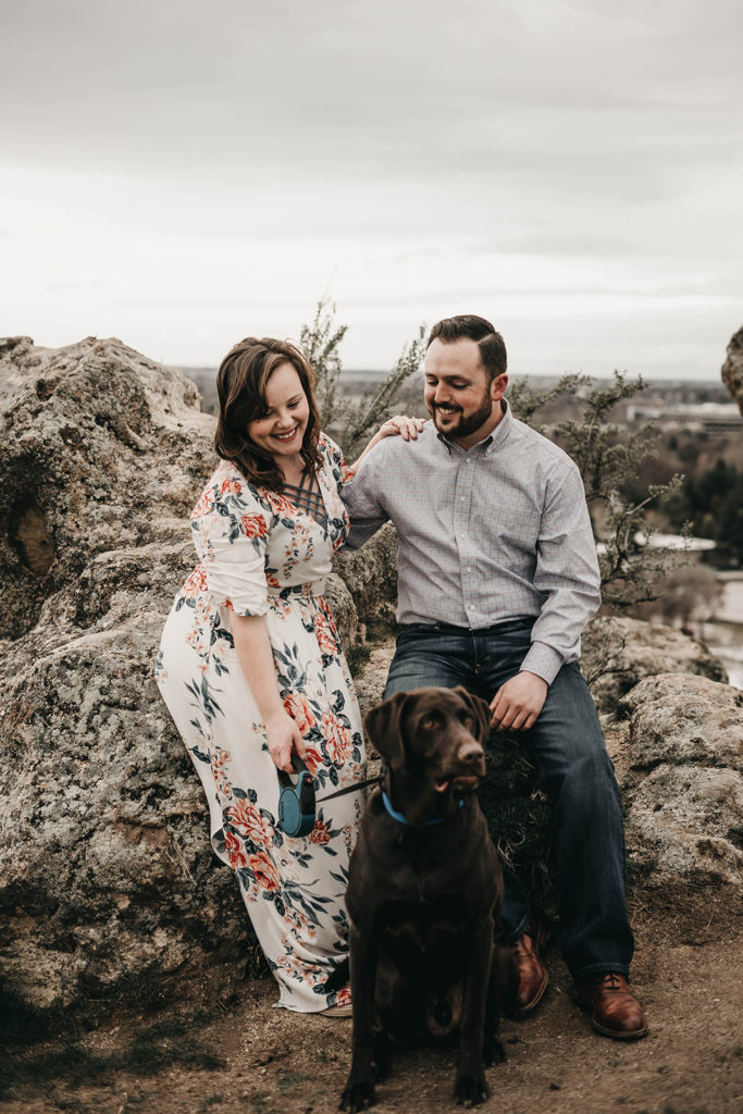couple smiles down at their chocolate lab during their engagement session at the military reserve