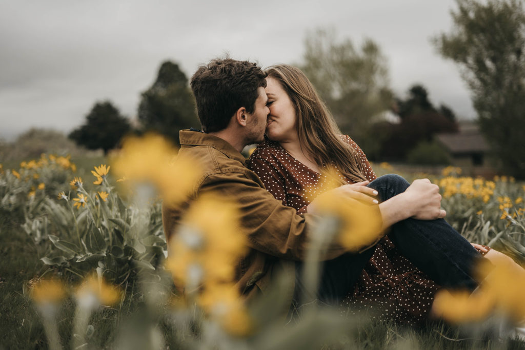 couple kisses as they sit among the balsamroot sunflowers in the boise foothills for their engagement session