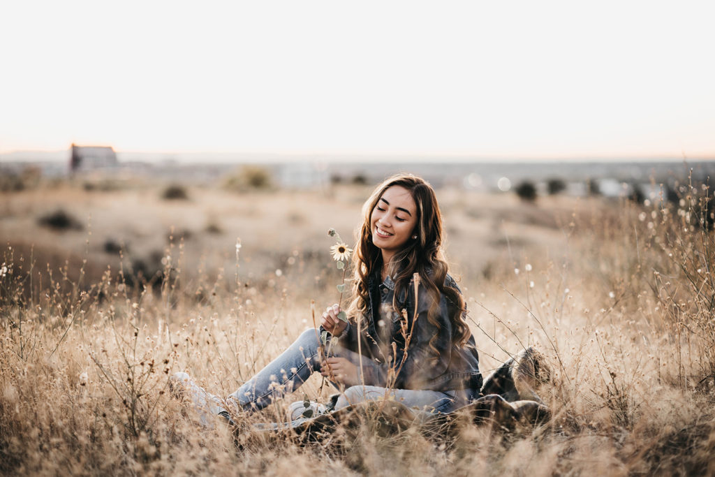 high school senior sits in the boise foothills during her senior session holding a balsamroot sunflower