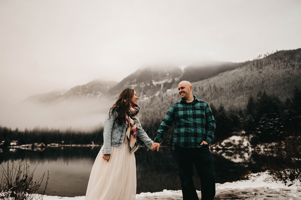 couple holds hands in front of a snowy gold creek pond for their snoqualmie pass engagement session
