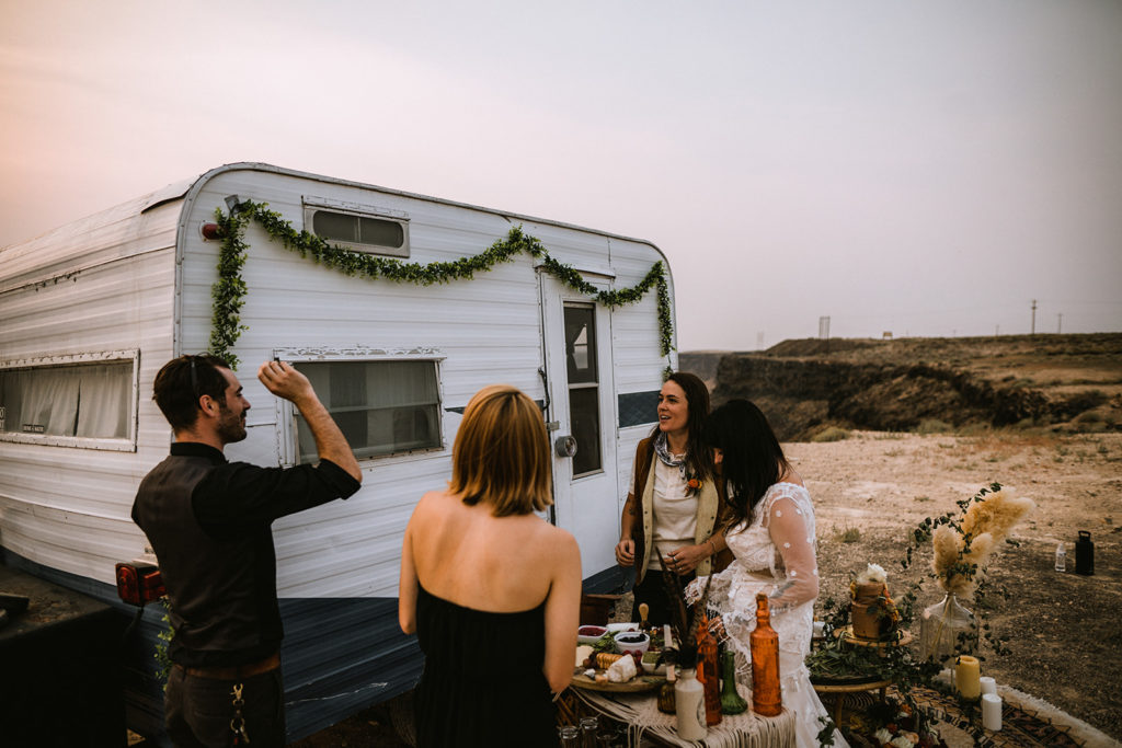 wedding guest stand in front of decorated vintage camper while enjoying charcuterie board after elopement ceremony