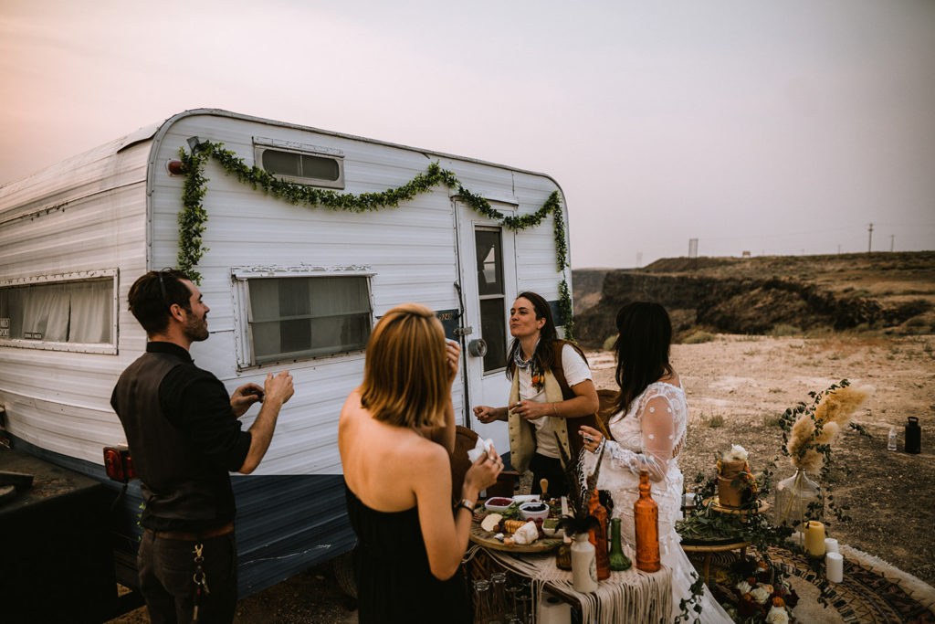 Elopement guests stand in front of the vintage camper trailer to eat from the charcuterie board