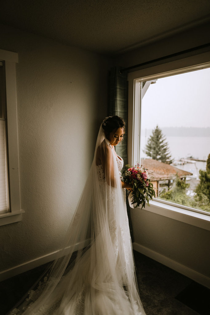 bride holding her bouquet and looking down at her veil in front of a window