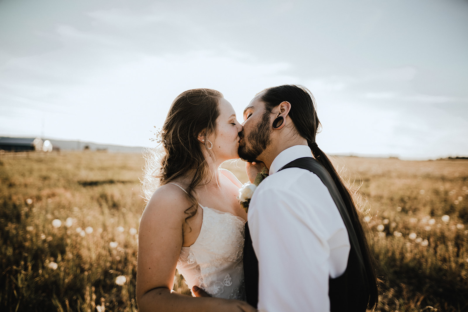 Bride and groom share a kiss in a pasture as the sun sets behind them.