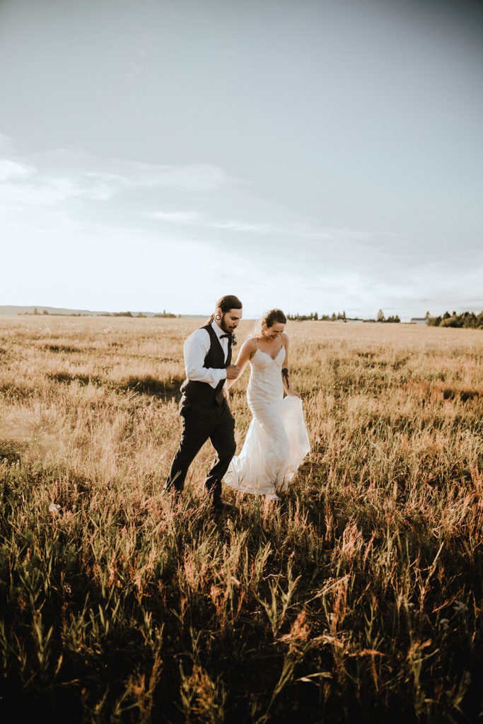 Bride and groom run through a pasture hand in hand as the sun sets in Donnelly Idaho