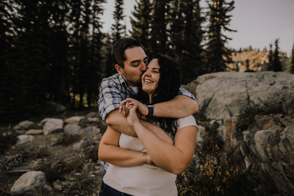 Couple embraces while man kisses his fiancee on the forehead during a blue lake engagement session in cascade, idaho