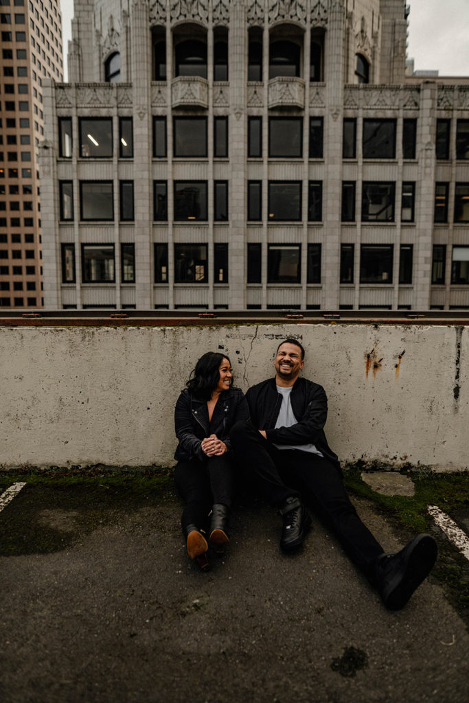 couple sitting down on a parking garage rooftop laughing during their engagement session in Downtown Seattle