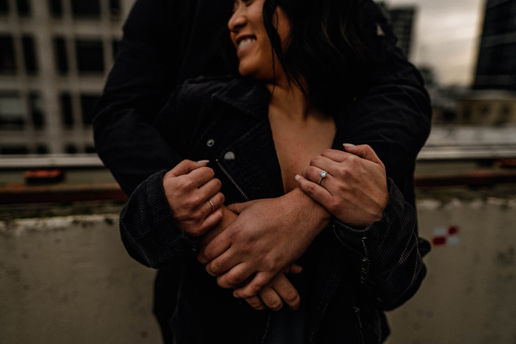 Couple stand on a rooftop of a parking garage holding each other and showing an engagement ring during their engagement session in Seattle