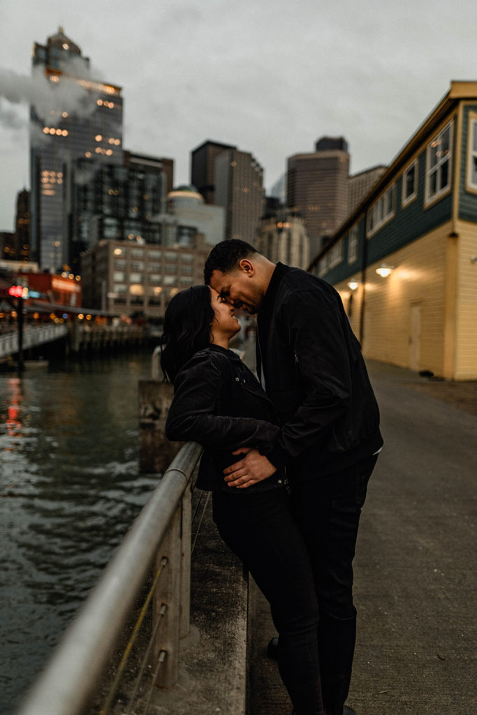 couple touches foreheads together as they lean on the railing of a pier in seattle