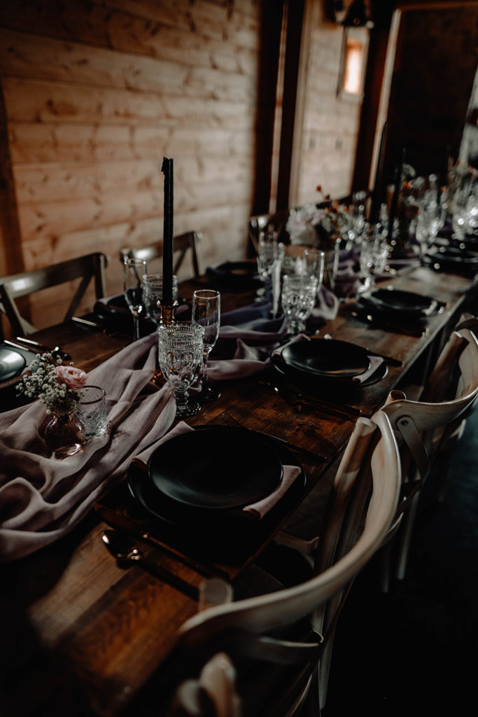 Detail shot of reception table decor, with black plates and chargers, crystal champagne and drink glasses, brass candlestick holders, and black candles on a dark wood table.