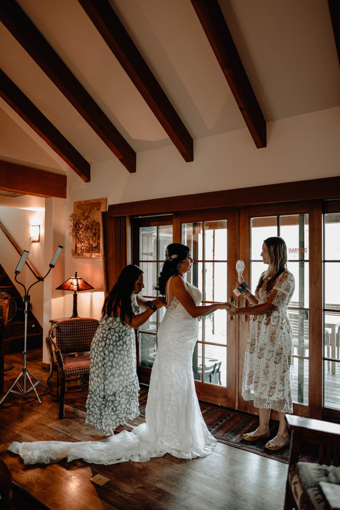 Bride gets into her wedding dress as she holds hands with her future sister-in-law in front of the cabin patio doors.