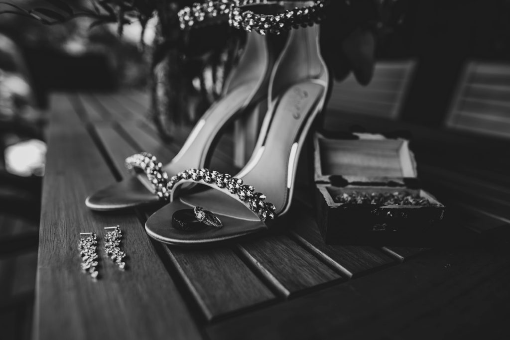 Black and white photo of bride and groom's rings sitting on bride's shoes with earrings in front