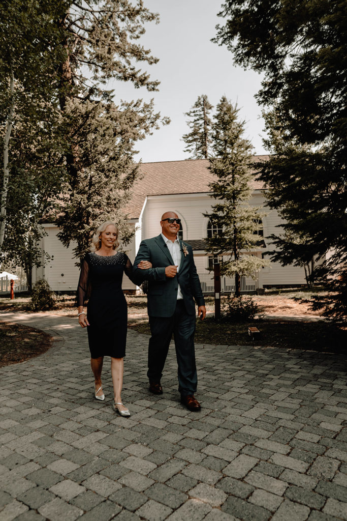 Groom walks with his mother down the aisle with the Tamarack chapel in the background