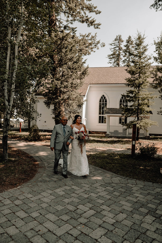 Bride and her father walk down the aisle with the Tamarack resort chapel in the background