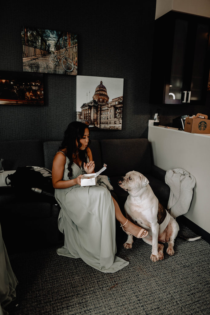 Bridesmaid sits on the couch and eats chicken nuggets as the bride's dog watches her eat in the getting ready suite
