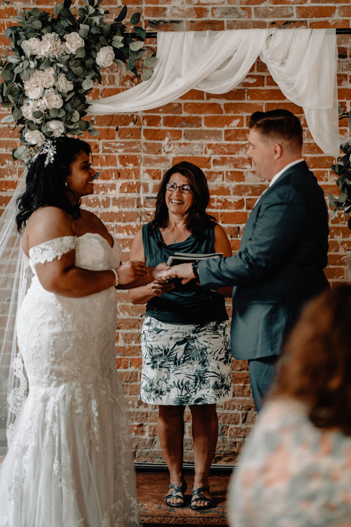 photo of the bride and groom looking at each other as they give each other their rings in front of the brick wall at beside bardenay