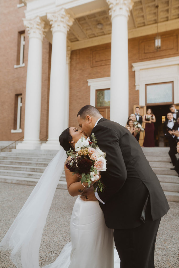 Newlyweds kiss after leaving the church as their wedding party stands behind them on the steps of St. Aloysius at Gonzaga