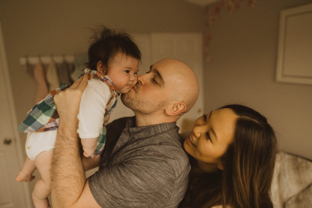 dad kisses his baby on the cheek while mom stands behind him in the baby's nursery